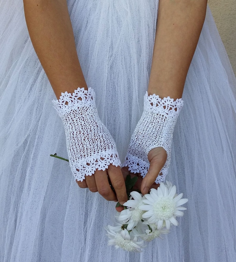 May include: A pair of white fingerless gloves featuring a delicate knit texture and intricate scalloped lace trim at the top and bottom edges. The gloves are crafted from a fine white thread and are worn over a white tulle fabric background.