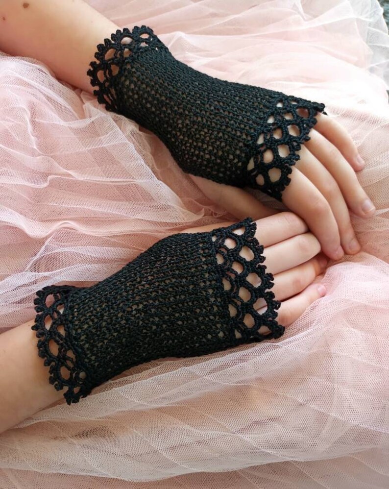 May include: Black crochet fingerless gloves with a scalloped edge. The gloves are made of a fine, open weave and are displayed on a pink tulle background. The gloves are a stylish accessory.