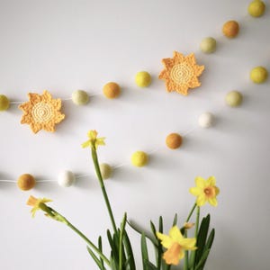 Op de afbeelding: Een decoratieve slinger met gele, oranje en witte vilten ballen geregen aan een wit koord. Twee gehaakte gele zonnen zijn afgewisseld tussen de vilten ballen. De slinger hangt tegen een witte muur, met narcissen in een vaas op de voorgrond.
