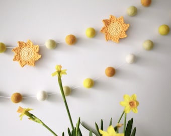 May include: A decorative garland featuring yellow, orange, and white felt balls strung on a white cord. Two crocheted yellow suns are interspersed among the felt balls. The garland is displayed against a white wall, with daffodils in a vase in the foreground.