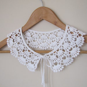 May include: White crocheted collar with a tie closure, displayed on a wooden hanger.