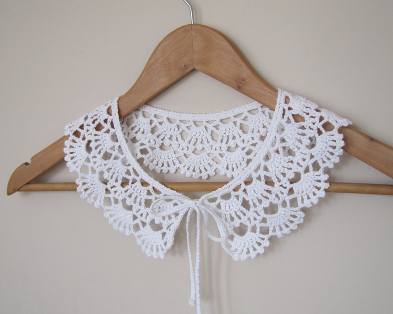 May include: White crocheted collar with a tie closure, displayed on a wooden hanger.