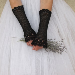 May include: A pair of black fingerless gloves featuring a fine knit texture and intricate scalloped lace trim at the cuffs. The gloves extend from the wrist to the mid-forearm, showcasing a delicate openwork pattern that contrasts with the solid black yarn.