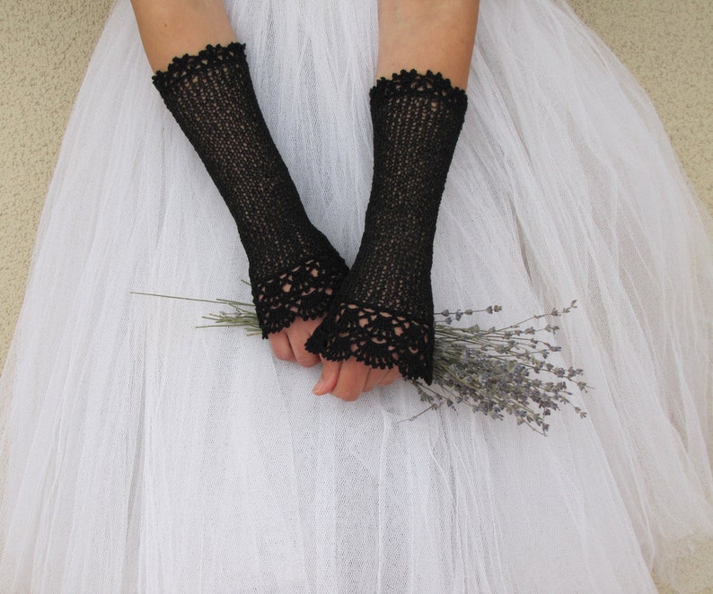 May include: A pair of black fingerless gloves featuring a fine knit texture and intricate scalloped lace trim at the cuffs. The gloves extend from the wrist to the mid-forearm, showcasing a delicate openwork pattern that contrasts with the solid black yarn.