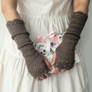 Può includere: Scaldamani lavorati a maglia marroni con bordo smerlato e motivo traforato decorativo. Gli scaldamani sono realizzati con un filato morbido e strutturato. La persona tiene in mano un libro a motivi floreali con un nastro rosa.
