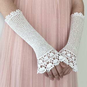 May include: A pair of white crochet fingerless gloves featuring a delicate openwork knit pattern and a scalloped lace edge at the wrist and forearm. The gloves extend from the knuckles to the mid-forearm, showcasing a fine, intricate thread construction.