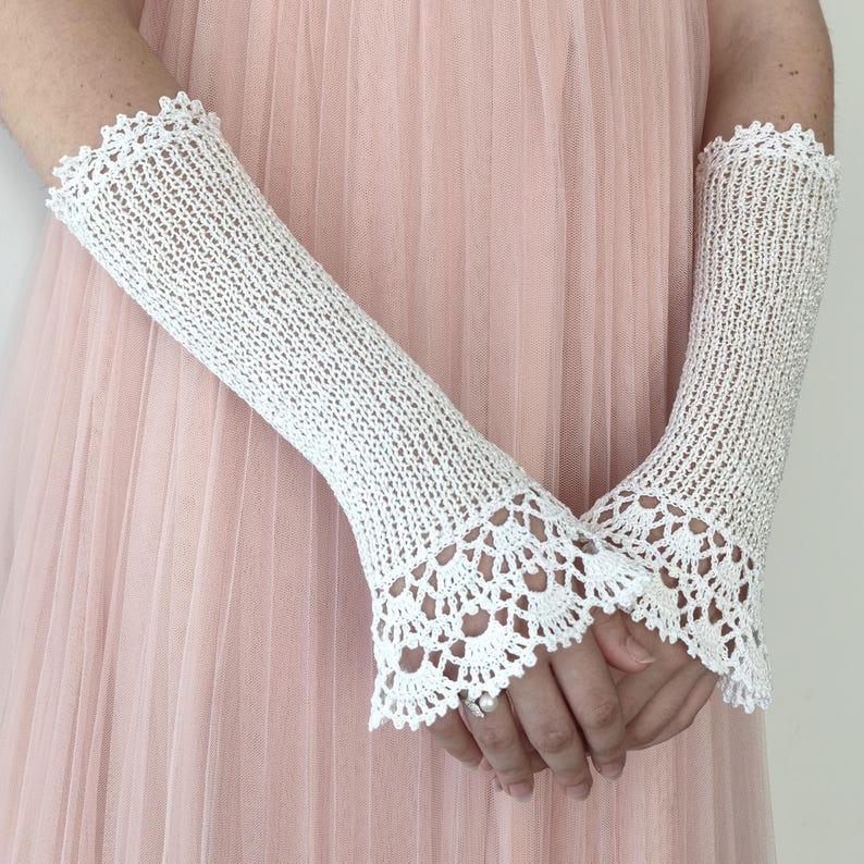 May include: A pair of white crochet fingerless gloves featuring a delicate openwork knit pattern and a scalloped lace edge at the wrist and forearm. The gloves extend from the knuckles to the mid-forearm, showcasing a fine, intricate thread construction.