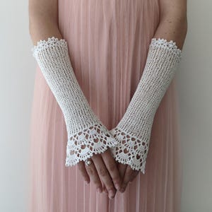 May include: A pair of off-white, elbow-length fingerless gloves crafted from a fine knit fabric. The gloves feature a delicate, intricate crochet lace trim at both the upper and lower edges, creating a scalloped pattern that extends over the wrists.