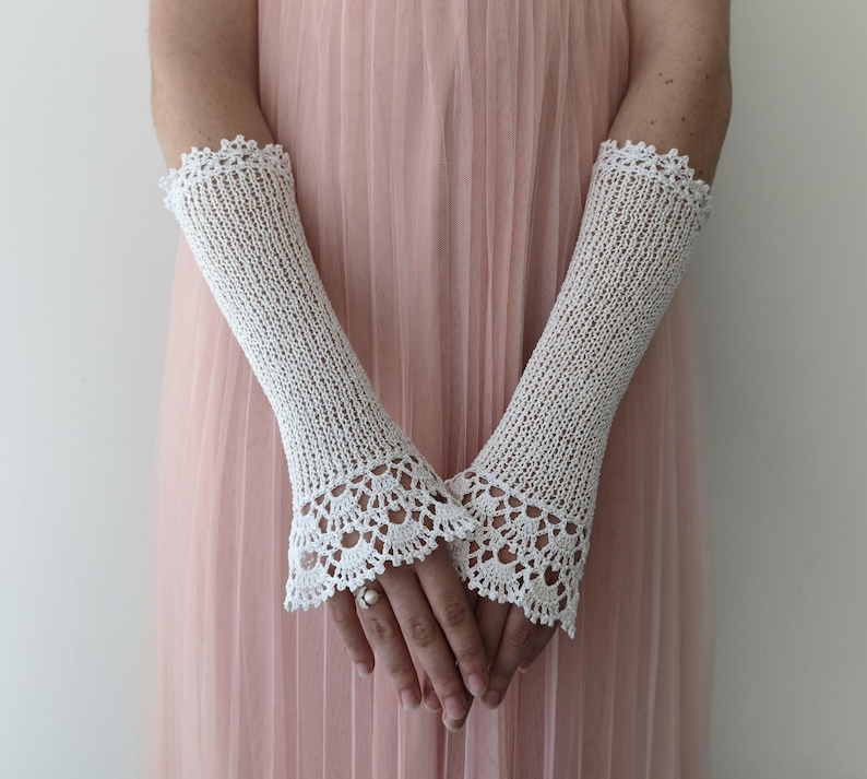 May include: A pair of off-white, elbow-length fingerless gloves crafted from a fine knit fabric. The gloves feature a delicate, intricate crochet lace trim at both the upper and lower edges, creating a scalloped pattern that extends over the wrists.
