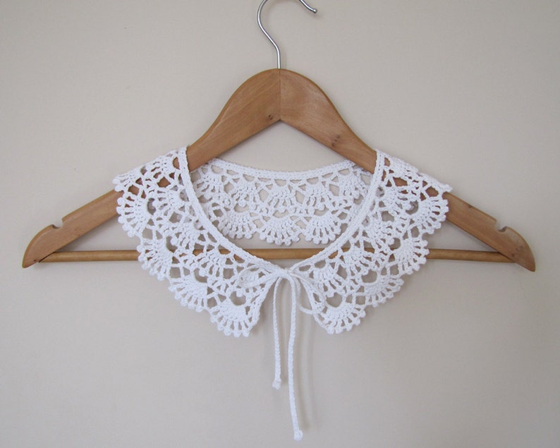 May include: White crocheted collar with a tie in the front, displayed on a wooden hanger.