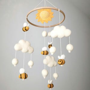 May include: A hanging mobile featuring a circular wooden frame with a yellow crocheted sun at the center. Suspended from the frame are white felted clouds, small white felted spheres, and yellow and black striped felted bees on white strings.