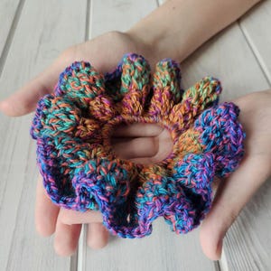 May include: A handmade, colorful crochet scrunchie. The scrunchie features a vibrant mix of blue, purple, orange, and green yarn. The scrunchie is held in open hands, showcasing its round shape and textured design. A stylish hair accessory.