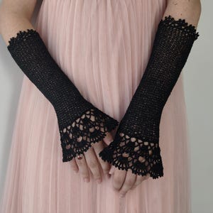 May include: A pair of black fingerless gloves crafted from a fine knit material, extending from the mid-forearm to the knuckles. The gloves feature a delicate, intricate crochet lace trim at the flared cuffs, adding texture to the design.