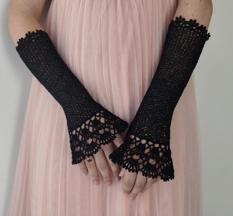 May include: A pair of black fingerless gloves crafted from a fine knit material, extending from the mid-forearm to the knuckles. The gloves feature a delicate, intricate crochet lace trim at the flared cuffs, adding texture to the design.