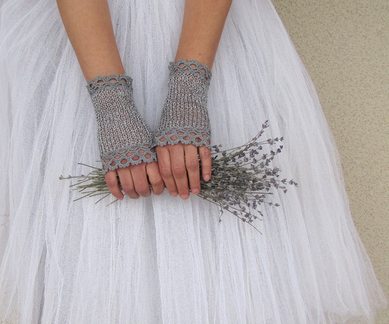 May include: A pair of gray fingerless gloves with a delicate lace trim. The gloves are knitted and held together with a bouquet of dried lavender. The gloves are a light gray color and are made of a soft, textured material.
