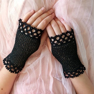 May include: Black fingerless gloves with a delicate, openwork crochet design. The gloves feature a scalloped edge at the wrist and around the fingers. The gloves are shown on a person's hands against a soft pink background.