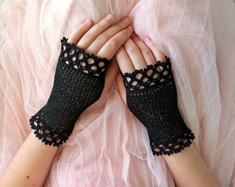 May include: Black fingerless gloves with a delicate, openwork crochet design. The gloves feature a scalloped edge at the wrist and around the fingers. The gloves are shown on a person's hands against a soft pink background.