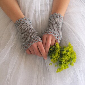 May include: A pair of light gray, fingerless knitted gloves with a scalloped edge. The gloves are worn over the hands, which are holding a small bouquet of yellow flowers. The background is a soft, white fabric.