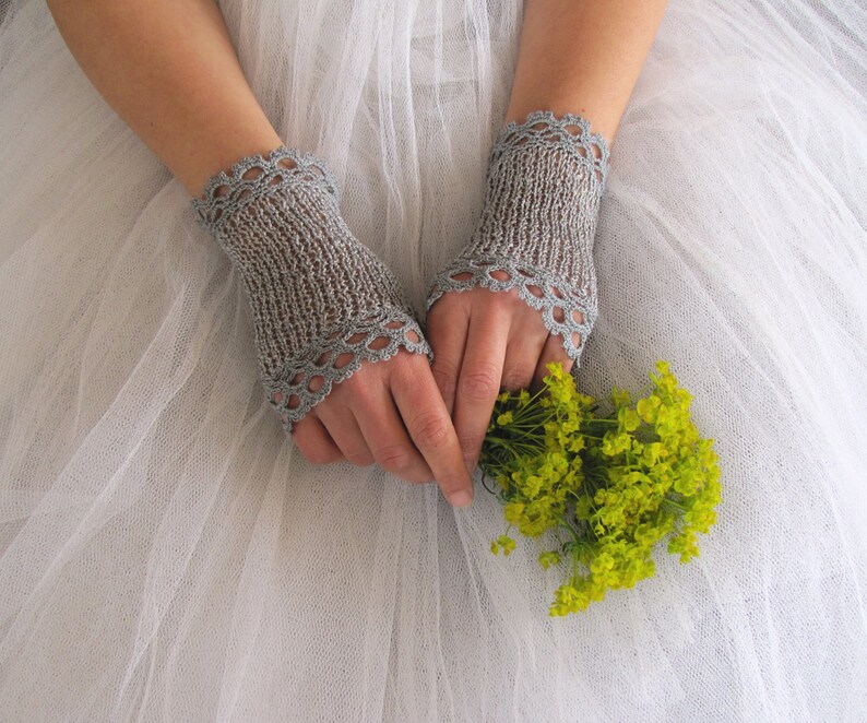 May include: A pair of light gray, fingerless knitted gloves with a scalloped edge. The gloves are worn over the hands, which are holding a small bouquet of yellow flowers. The background is a soft, white fabric.