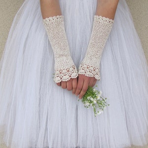 May include: A pair of off-white, elbow-length fingerless gloves featuring a delicate, intricate crochet lace trim at the cuffs and a fine knit texture throughout the main body, worn over a white tulle skirt while holding a small bouquet of white flowers.