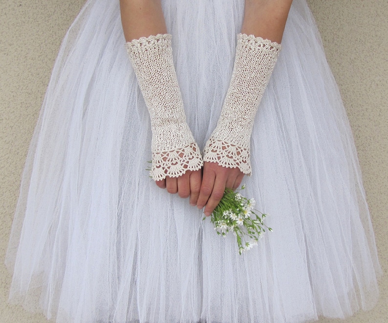 May include: A pair of off-white, elbow-length fingerless gloves featuring a delicate, intricate crochet lace trim at the cuffs and a fine knit texture throughout the main body, worn over a white tulle skirt while holding a small bouquet of white flowers.
