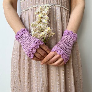 May include: A pair of light purple fingerless gloves featuring a delicate openwork knit pattern and a scalloped crochet edge. The gloves are worn on the wrists and extend to the knuckles, showcasing a textured yarn construction in a soft lavender hue.