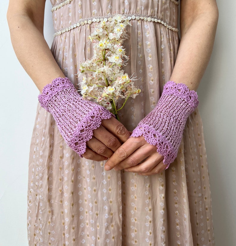 May include: A pair of light purple fingerless gloves featuring a delicate openwork knit pattern and a scalloped crochet edge. The gloves are worn on the wrists and extend to the knuckles, showcasing a textured yarn construction in a soft lavender hue.