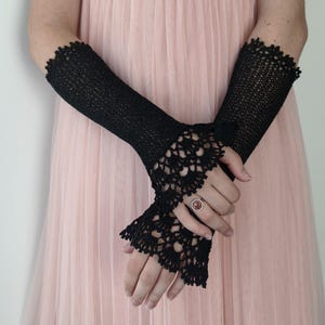 May include: A pair of black fingerless gloves featuring a fine knit texture on the forearm and an intricate, scalloped crochet lace pattern at the wrist. The gloves extend from the mid-forearm to the base of the fingers, showcasing detailed craftsmanship.