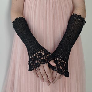 May include: A pair of black fingerless gloves featuring a fine crochet knit pattern throughout the length of the arm. The gloves are finished with an intricate, scalloped lace-like crochet border at the wrist, extending over the back of the hand.