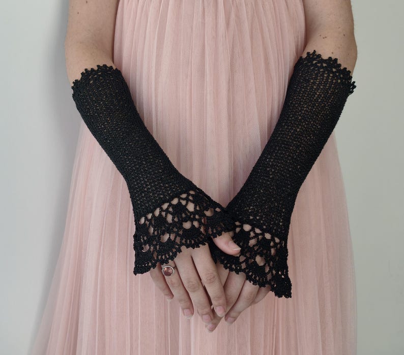 May include: A pair of black fingerless gloves featuring a fine crochet knit pattern throughout the length of the arm. The gloves are finished with an intricate, scalloped lace-like crochet border at the wrist, extending over the back of the hand.