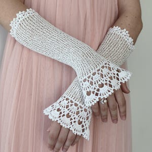 May include: A pair of white crochet fingerless gloves featuring an intricate openwork knit pattern throughout the forearm and a decorative scalloped lace edge at the wrist. The gloves are crafted from fine white thread with a delicate texture.