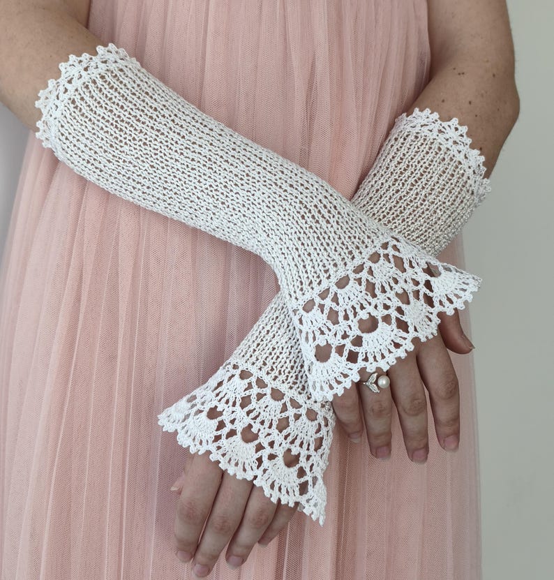 May include: A pair of white crochet fingerless gloves featuring an intricate openwork knit pattern throughout the forearm and a decorative scalloped lace edge at the wrist. The gloves are crafted from fine white thread with a delicate texture.