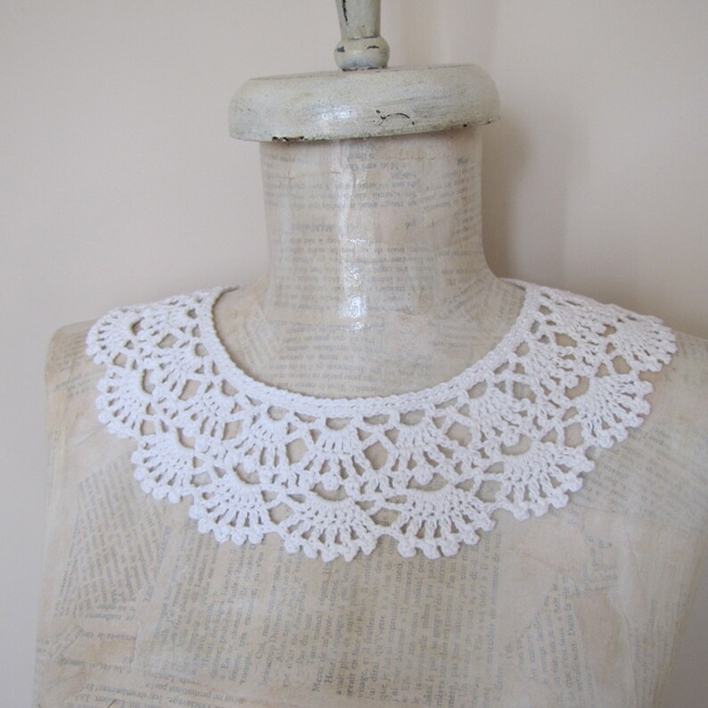 May include: White crocheted collar on a white mannequin. The collar is made of delicate lace and has a scalloped edge.