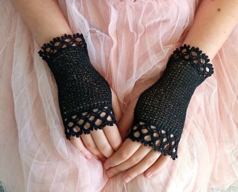 May include: A pair of black fingerless gloves. The gloves are made of a delicate, open-weave material. The cuffs and the top edge of the gloves are adorned with a scalloped, lace-like trim. The gloves are designed to cover the hand up to the knuckles.