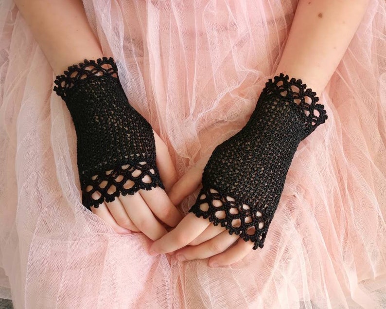 May include: Black fingerless gloves with a delicate, openwork design. The gloves have a scalloped edge and are made of a fine, knitted material. The gloves are shown against a soft pink background.