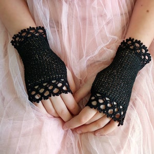 May include: A pair of black, fingerless crochet gloves. The gloves have a delicate, openwork design with a scalloped edge. The gloves are made of a fine, dark yarn and are displayed against a soft, pink background.