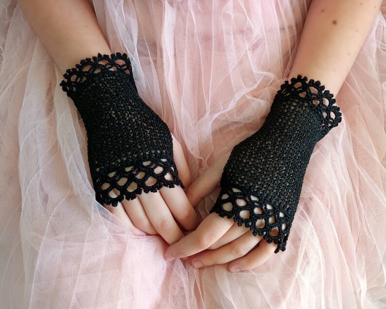 May include: A pair of black, fingerless crochet gloves. The gloves have a delicate, openwork design with a scalloped edge. The gloves are made of a fine, dark yarn and are displayed against a soft, pink background.