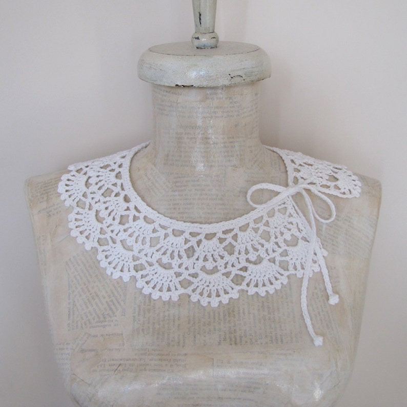 May include: White crocheted collar with a delicate lace pattern. The collar is tied at the back with a white ribbon.