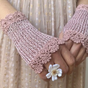 May include: A pair of dusty pink crochet fingerless gloves featuring a delicate scalloped edge pattern at the wrists and knuckles. A white flower-shaped ring with a gold-toned center is worn on one finger, complementing the intricate knit texture.