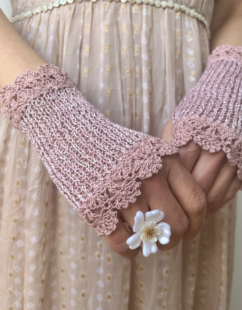 May include: A pair of dusty pink crochet fingerless gloves featuring a delicate scalloped edge pattern at the wrists and knuckles. A white flower-shaped ring with a gold-toned center is worn on one finger, complementing the intricate knit texture.