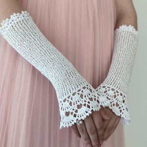 May include: A pair of white fingerless gloves featuring a delicate knit texture and intricate scalloped crochet lace trim at the top and bottom edges. The gloves extend from the wrist to the mid-forearm, showcasing a detailed openwork pattern.