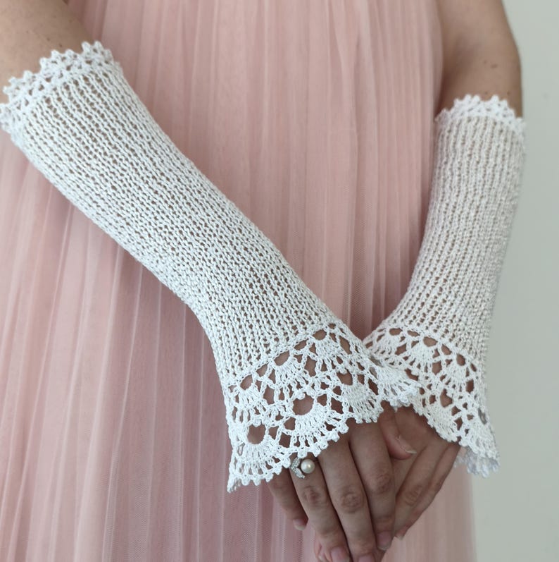 May include: A pair of white fingerless gloves featuring a delicate knit texture and intricate scalloped crochet lace trim at the top and bottom edges. The gloves extend from the wrist to the mid-forearm, showcasing a detailed openwork pattern.