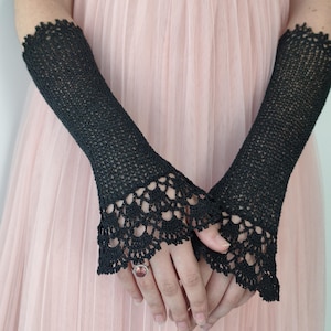 May include: A pair of black fingerless gloves featuring a fine knit texture and an intricate, scalloped crochet lace trim at the wrists. The gloves extend up the forearms and are worn over a pale pink dress with a pleated, layered fabric texture.