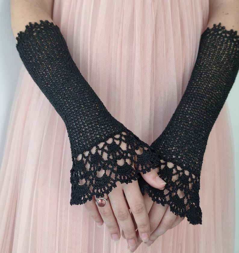 May include: A pair of black fingerless gloves featuring a fine knit texture and an intricate, scalloped crochet lace trim at the wrists. The gloves extend up the forearms and are worn over a pale pink dress with a pleated, layered fabric texture.