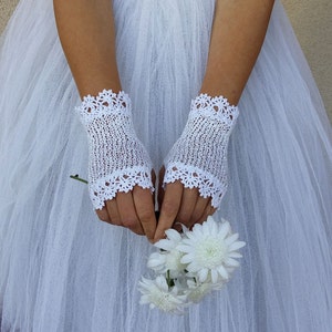 May include: A pair of white fingerless gloves featuring a delicate crochet knit pattern with scalloped edges at the top and bottom. The gloves are crafted from a fine white thread and are worn over the hands, which are holding a small bouquet of white flowers.