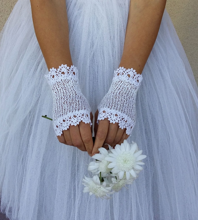 May include: A pair of white fingerless gloves featuring a delicate crochet knit pattern with scalloped edges at the top and bottom. The gloves are crafted from a fine white thread and are worn over the hands, which are holding a small bouquet of white flowers.