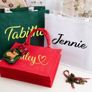 K&ouml;nnte beinhalten: Drei rechteckige Jute-Tragetaschen in Gr&uuml;n, Rot und Wei&szlig;. Die gr&uuml;ne Tasche tr&auml;gt den Namen Tabitha in Gelb, die wei&szlig;e Tasche den Namen Jennie in Schwarz und die rote Tasche den Namen Haley in Gelb mit einem Herzsymbol.
