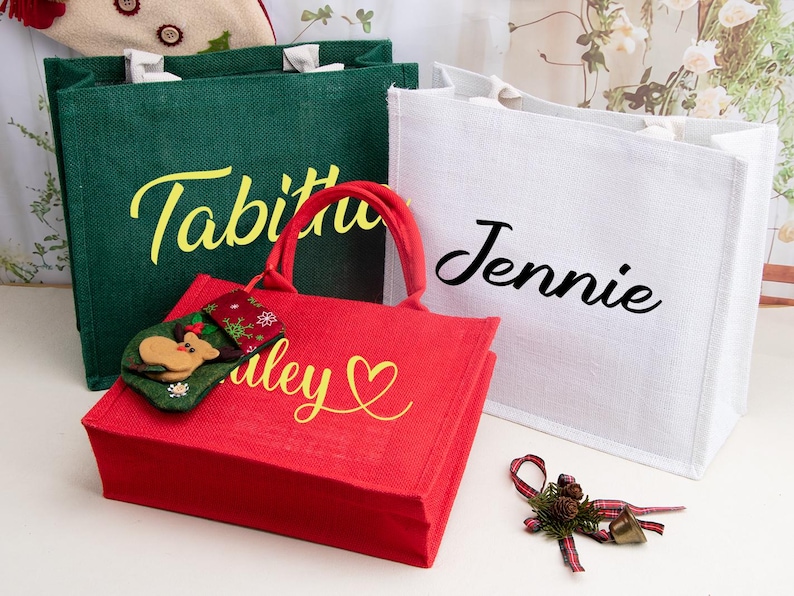 K&ouml;nnte beinhalten: Drei rechteckige Jute-Tragetaschen in Gr&uuml;n, Rot und Wei&szlig;. Die gr&uuml;ne Tasche tr&auml;gt den Namen Tabitha in Gelb, die wei&szlig;e Tasche den Namen Jennie in Schwarz und die rote Tasche den Namen Haley in Gelb mit einem Herzsymbol.