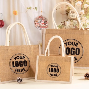 Könnte beinhalten: Drei Jute-Tragetaschen in verschiedenen Größen mit weißen Griffen und Verzierungen. Jede Tasche hat ein schwarzes kreisförmiges Logo mit der Aufschrift "YOUR LOGO HERE". Die Taschen werden auf einer weißen Oberfläche mit festlichen Dekorationen im Hintergrund präsentiert.