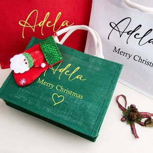 K&ouml;nnte beinhalten: Drei Jute-Tragetaschen in Rot, Gr&uuml;n und Wei&szlig; mit wei&szlig;en Henkeln. Die Taschen tragen den Namen Adela in Schreibschrift, dazu der Text Merry Christmas und ein Herzsymbol auf der gr&uuml;nen Tasche. Ein kleiner Weihnachtsstiefel und eine Schleife liegen bei.